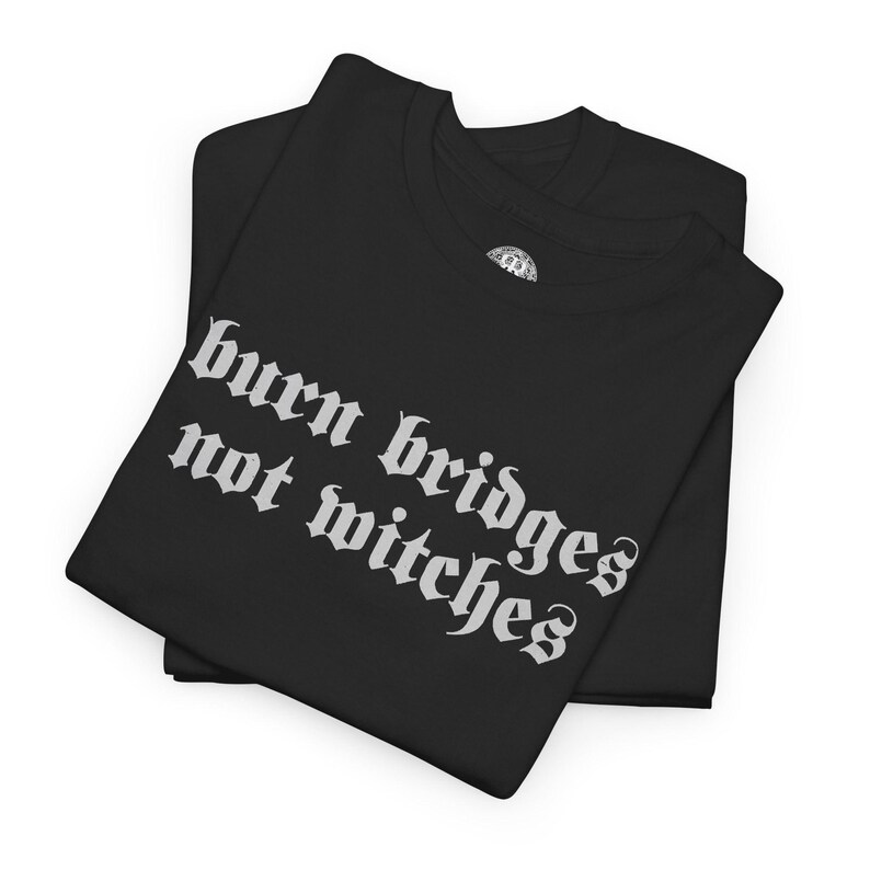 May include: A black t-shirt featuring the white text "burn bridges not witches" printed in a gothic-style font across the front. The shirt is folded, showing a short-sleeved design with a round neckline and a small circular logo on the inner collar.