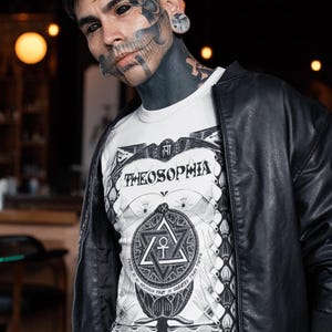 May include: A man with black and white tattoos, including a skull on his face, is wearing a white t-shirt with a black and white graphic design that says "Theosophia" and a black leather jacket.