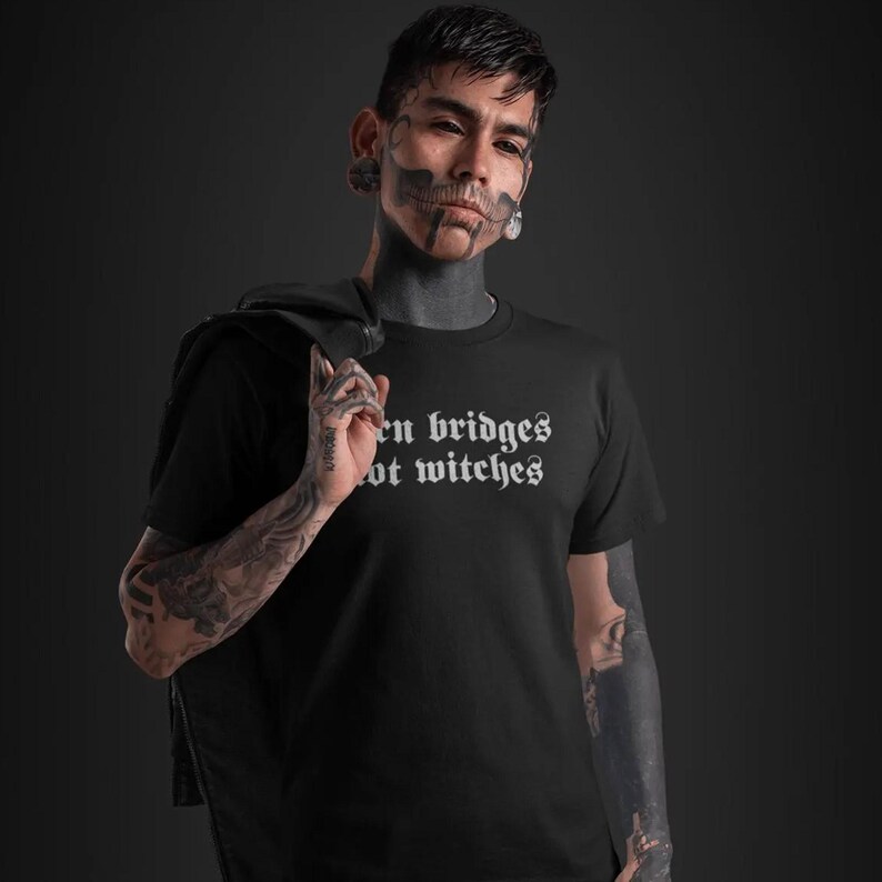 May include: A black short-sleeved t-shirt featuring white text printed on the chest that reads "burn bridges not witches" in a gothic-style font. The garment is made of a solid black fabric and has a standard crew neckline and short sleeves.