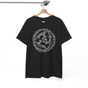 May include: A black short-sleeve t-shirt featuring a white circular graphic of three hares arranged in a triangle formation, surrounded by a detailed leaf and vine border. The design includes a small triquetra symbol in the center.