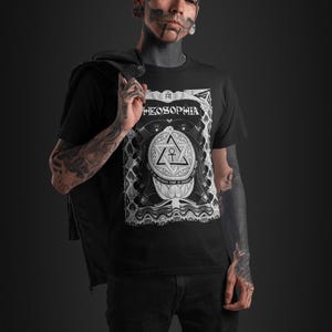 May include: A black t-shirt with a white graphic design featuring a stylized triangle with an ankh symbol inside. The text "THEOSOPHIA" is above the triangle. The graphic is surrounded by a decorative border.