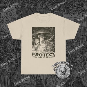 May include: Cream-colored t-shirt featuring a black and white illustration of a toad, mushroom, and small creature in a forest scene. The word "PROTECT" is printed above "LITTLE LIVES OF THE WOODS". A skull emblem is visible at the bottom right.