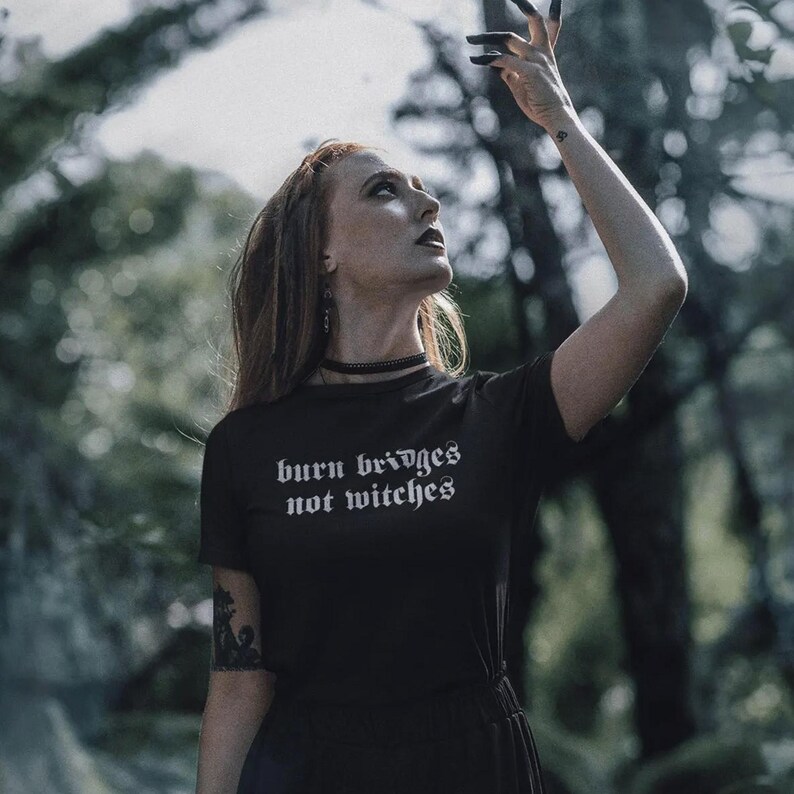 May include: A black short-sleeved t-shirt featuring the white text "burn bridges not witches" in a gothic-style font. The shirt has a crew neckline and is worn with a thin black choker necklace, showcasing a dark aesthetic design.