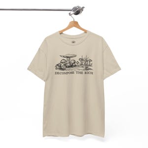 May include: Beige t-shirt with a black and white illustration of mushrooms and the text "DECOMPOSE THE RICH". The shirt is hanging on a wooden hanger.