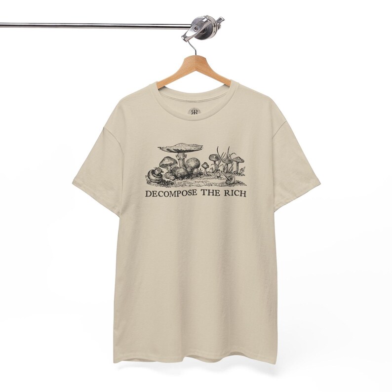 May include: Beige t-shirt with a black and white illustration of mushrooms and the text "DECOMPOSE THE RICH". The shirt is hanging on a wooden hanger.