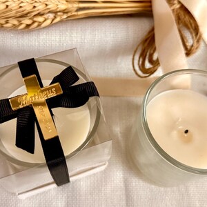 May include: Two glass votive candles, one in a clear box with a black ribbon and gold cross. The cross has the name "Matheus" and the date "16-07-2023". The other candle is in a clear glass. Wheat stalks and ribbon are in the background.