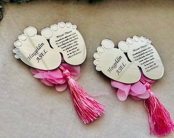 May include: Two silver baby foot-shaped plaques with pink fabric flowers and tassels. The plaques have the text "Nazar Duası" and "Hoşgeldin ASEL" on them.
