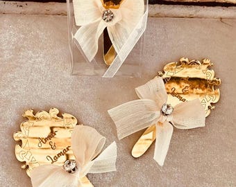May include: Gold-colored wedding favors with a decorative bow and a small jewel in the center. The favors have the names "Amar & Lamar" engraved on them. One favor is packaged in a clear box.