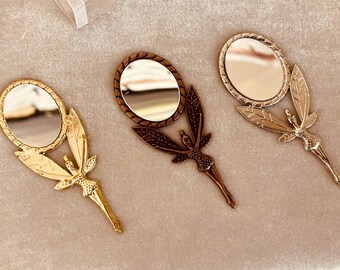 May include: Three ornate hand mirrors with fairy-like designs. Each mirror has a different color: gold, silver, and bronze.