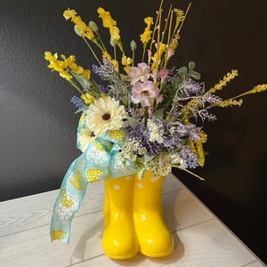 May include: Yellow wellies with white polka dots, filled with a bouquet of yellow, purple, and white flowers, and a blue ribbon with white flowers.