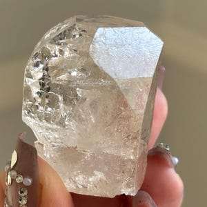 May include: A clear quartz crystal with a faceted top and a rough, textured body. The crystal is held in a hand, showcasing its natural, raw beauty. The crystal's color is a pale, translucent beige.
