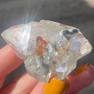 May include: A clear quartz crystal cluster with inclusions of black and orange minerals. The crystal is being held in a hand with yellow nail polish.