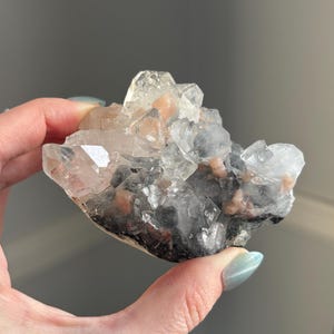 May include: A cluster of clear quartz crystals with a pink and gray hue. The crystals are clustered together in a natural formation.