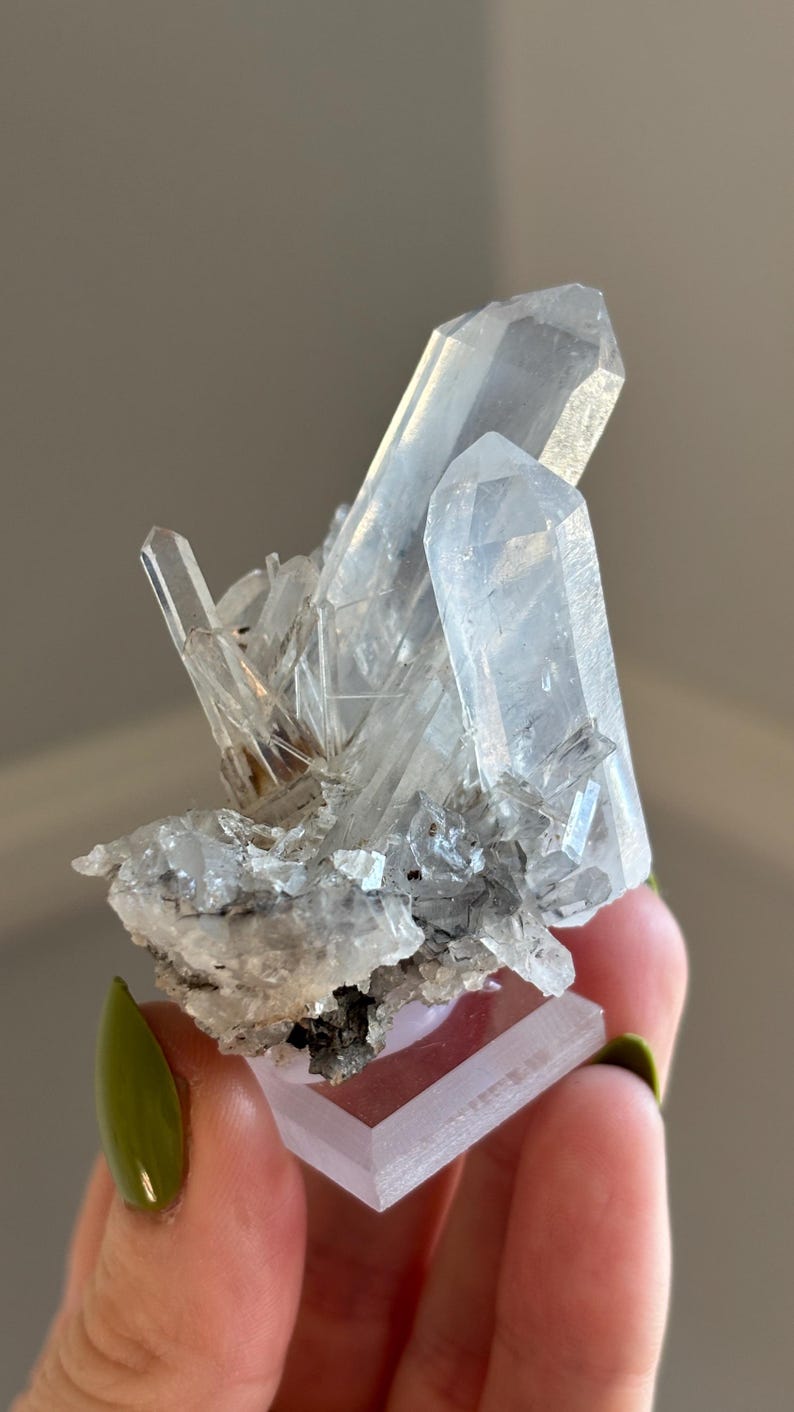 May include: A cluster of clear quartz crystals, of varying sizes and shapes, with a light grey and white base. The crystals are translucent, reflecting light and showcasing their natural formations. The quartz sits on a rectangular, light pink base.