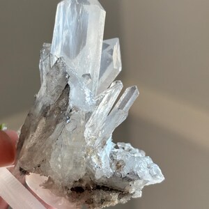 May include: A cluster of clear quartz crystals with sharp, pointed terminations. The crystals have a translucent appearance with some areas of grey and black inclusions. The quartz is held by a white base.
