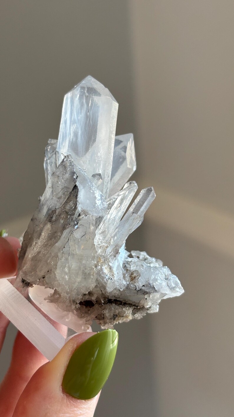 May include: A cluster of clear quartz crystals with sharp, pointed terminations. The crystals have a translucent appearance with some areas of grey and black inclusions. The quartz is held by a white base.