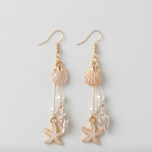 May include: A pair of gold-tone seashell earrings with pearl and starfish accents. The earrings feature a seashell at the top, a string of pearls, and a starfish charm. The earrings are designed with a beach or ocean theme.