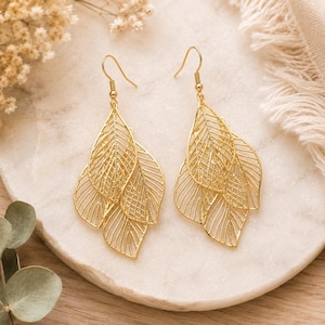 May include: A pair of gold-colored leaf earrings. Each earring features three layered, openwork leaf shapes with intricate vein detailing. The earrings hang from gold-colored ear wires, showcasing a delicate and elegant design.