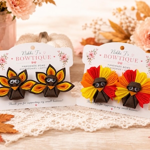 May include: Two white display cards labeled Nikki T's BOWTIQUE, each holding two turkey-shaped hair bows. The left card features brown, orange, and yellow ribbon turkeys. The right card displays turkeys with red, orange, and yellow ribbon feathers.