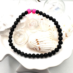 Könnte beinhalten: Schwarzes Lava-Stein-Perlenarmband mit rosa Akzenten. Das Armband besteht aus kleinen, runden, schwarzen Perlen, die aneinandergereiht sind. Der Verschluss ist leuchtend rosa. Das Armband wird auf einer weißen Oberfläche mit Muscheln präsentiert.
