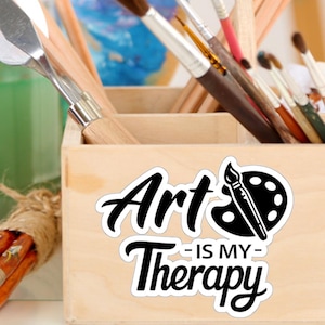Könnte beinhalten: Eine Holzkiste mit Pinseln und einem Aufkleber mit der Aufschrift "Art is my Therapy" mit einem Pinsel- und Paletten-Design.