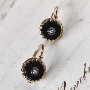 Puede incluir: Un par de pendientes dorados con centros circulares negros y una pequeña gema de color claro en el centro. Los pendientes tienen un borde decorativo y texturizado y un cierre de gancho. El fondo es una superficie clara y estampada.