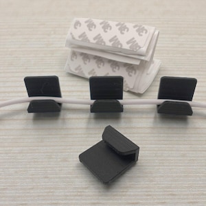 May include: Black cable clips with adhesive backing, holding a white cable. Several clips are attached to the cable, while one is separate. A stack of adhesive pads is in the background. The clips are designed to organize and secure cables.