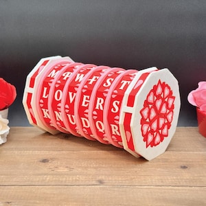 May include: A wooden puzzle with red and white accents. The puzzle has rotating rings with the word "LOVERS" spelled out. The end of the puzzle features a heart design. The puzzle is on a wooden surface.