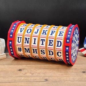 May include: A cylindrical rotating puzzle toy featuring six white rings with blue letters and symbols. The rings spell UNITED and #MHSDC. The end caps are red with blue borders and white star patterns. The structure is made of red, white, and blue components.