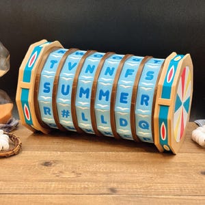 May include: A cylindrical puzzle game featuring rotating light blue segments with dark blue letters and symbols. The device has brown structural rings and tan end caps decorated with colourful geometric patterns. The letters spell out SUMMER.