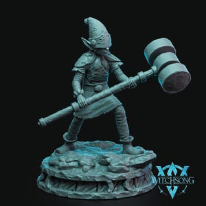 Puede incluir: Una figurita miniatura pintada de gris de una criatura fantástica con orejas puntiagudas, que lleva un casco y sostiene un martillo grande. La figura está de pie sobre una base rocosa con el texto "WITCHSONG" impreso en la base.