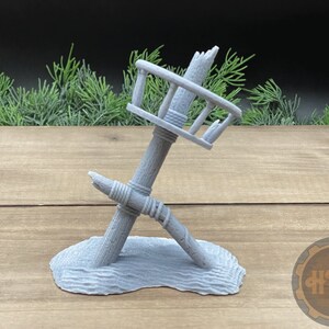 以下が含まれることがあります： グレーの3Dプリントされた、破損した木製の監視塔のミニチュア。手すり付き。塔は横に傾いており、グレーのテクスチャのあるベースの上に置かれています