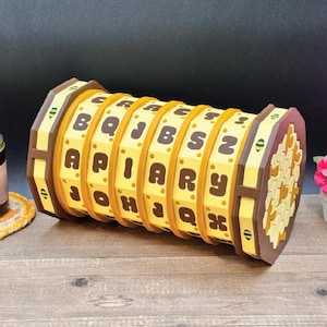 Puede incluir: Un rompecabezas de madera con cilindros giratorios con letras que deletrean "APIARY". El rompecabezas es amarillo y marrón, con un diseño de panal en un extremo. Las letras están en una fuente marrón oscuro.