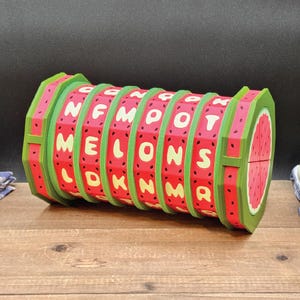 May include: A cylindrical puzzle toy featuring a watermelon theme with green edges and red segments decorated with black seeds. The rotating rings display cream-coloured letters that spell out MELONS, with additional letters visible on the surface.