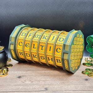May include: A green and gold Celtic-themed puzzle box with rotating lettered cylinders. The cylinders spell out "CELTIC" and other letters. The box has intricate Celtic knot designs on the ends and is set on a wooden surface.