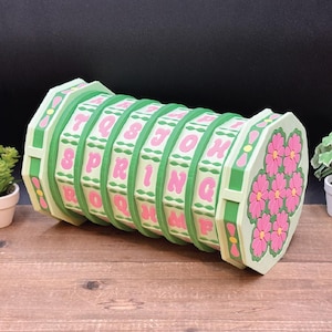 Puede incluir: Un rompecabezas decorativo verde claro y rosa con la palabra "SPRING" en letras rosas. El rompecabezas tiene forma cilíndrica con diseños de flores en los extremos. El rompecabezas está hecho de plástico y se encuentra sobre una superficie de madera.