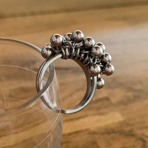 Peut inclure: Une bague en argent avec un groupe de petites boules argentées. La bague est faite de fil et les boules sont attachées au fil. La bague est posée sur une surface en verre.