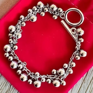 Peut inclure: Un bracelet argenté avec un fermoir à glissière et plusieurs rangées de perles rondes. Le bracelet est présenté sur un fond en tissu rose vif.