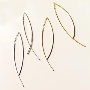 Op de afbeelding: Drie paar delicate, minimalistische oorbellen. Twee paar zijn zilver en één paar is goud. Elke oorbel is een dunne, gebogen draad met een klein kettinkje aan de bovenkant.