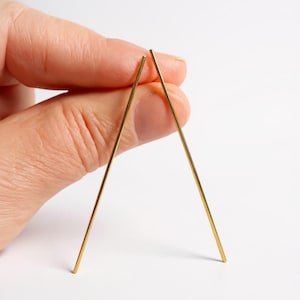 Op de afbeelding: Twee goudkleurige, rechte oorringen tegen een witte achtergrond. De oorbellen zijn dun en lijken van metaal te zijn. De oorbellen zijn onder een hoek geplaatst om een V-vorm te creëren.
