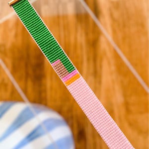 Peut inclure: Un bracelet de perles avec un fermoir doré. Le bracelet présente un motif de perles vertes, roses, marron et orange. L'autre moitié du bracelet est rose. Le bracelet est sur une surface en bois.