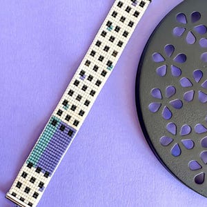 Peut inclure: Un bracelet de perles avec une base blanche, présentant un motif de carrés noirs. Le bracelet a une section de perles turquoise et violettes. Le bracelet est fait de petites perles et possède un fermoir et une chaîne argentés.