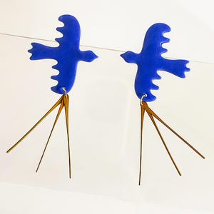 Pode incluir: Um par de brincos em forma de pássaro azul cobalto com pingentes lineares dourados. Os brincos são exibidos em um fundo branco. Os pássaros têm um design estilizado e recortado com formas detalhadas de asas e cauda.
