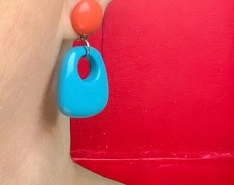 Peut inclure: Gros plan sur une oreille portant une paire de boucles d'oreilles rétro. Les boucles d'oreilles sont composées d'un petit clou orange et d'une plus grande breloque turquoise en forme de goutte d'eau avec une découpe circulaire. Le fond est rouge uni.