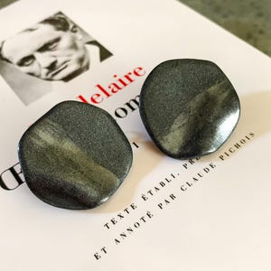 Peut inclure: Deux boucles d'oreilles clous de couleur gris foncé, de forme irrégulière, avec un éclat métallique. Les boucles d'oreilles sont présentées sur une surface blanche avec du texte et une image en noir et blanc d'une personne.