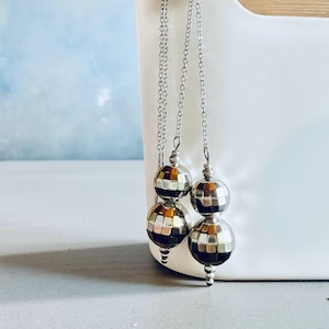 Può includere: Orecchini color argento con un design a sfera da discoteca. Ogni orecchino presenta due perline sferiche a specchio sospese da una delicata catena. Gli orecchini sono appesi davanti a un contenitore bianco con un accento in legno, su uno sfondo azzurro.
