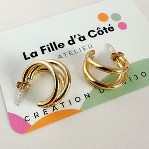 Peut inclure: Une paire de boucles d'oreilles créoles dorées avec un design à trois anneaux. Les boucles d'oreilles sont présentées sur une carte blanche avec le texte "La Fille d'à Côté ATELIER CREATION". Elles ont une finition brillante.
