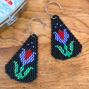 May include: Black beaded earrings with a floral design. Each earring has a black background with a red, blue, and green flower. The earrings have silver ear wires and a triangular shape.