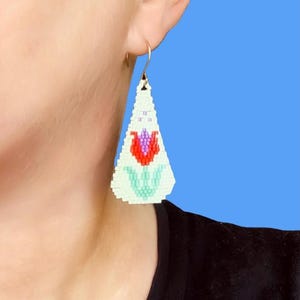 May include: A beaded earring with a gold hook. The earring is a white, triangular shape with a red, purple, and green floral design. The earring is worn on a person's ear.
