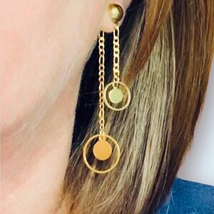 Könnte beinhalten: Goldfarbene Hängeohrringe. Jeder Ohrring hat einen kleinen, runden Stecker, eine Kette, einen kreisförmigen Rahmen und eine farbige Scheibe. Die Scheiben sind orange und grün. Die Ohrringe werden am Ohr getragen.